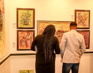 A woman and man viewing a collection of framed African paintings in a warmly lit gallery. The artworks feature expressive brushstrokes, vibrant colors, and emotionally rich scenes, reflecting rare themes and deep cultural meaning—similar to the pieces Cheryl discovered at Ovico Gallery. 
